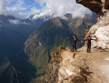 Two people standing on a cliff edge in front of a mountain view